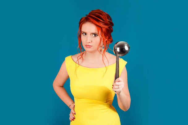 Angry upset frustrated woman holding ladle, dipper big spoon threatening someone. Caucasian person in yellow dress retro vintage redhead hairstyle on blue studio background. Parenting and relationship - Photo, Image