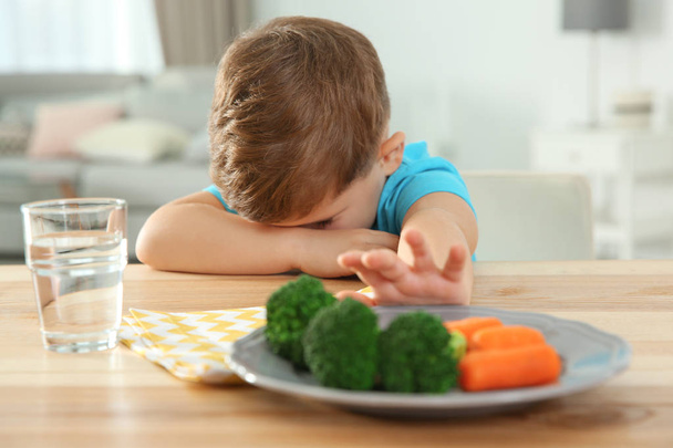 Sad little boy eating vegetable salad at table in room - Photo, image