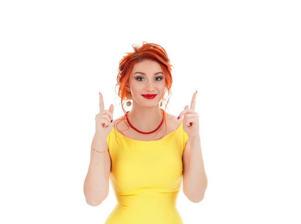 Close-up portrait of surprised pretty young woman in yellow dress pointing with two fingers up, looking at camera smiling, isolated on white studio background. Caucasian girl with retro hairstyle - Фото, зображення