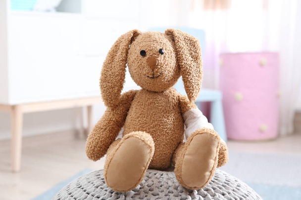 Toy bunny with bandaged pad on pouf indoors. Children's doctor - Φωτογραφία, εικόνα