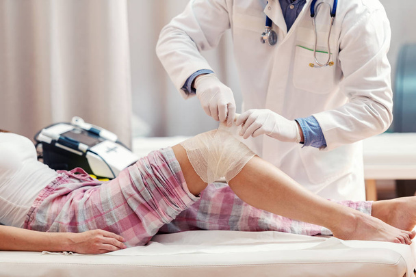 Zblízka pacientka v pyžamu vleže v nemocniční posteli, zatímco lékař v bílé uniformě skládací zraněné koleno. - Fotografie, Obrázek