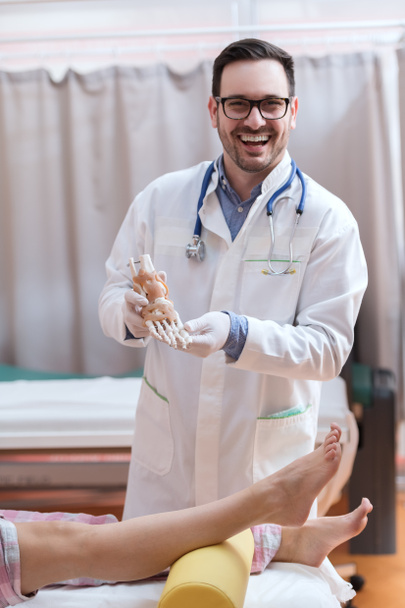 Smiling doctor in white uniform holding foot model while standing in hospital. In front of him patient's legs. - Photo, Image