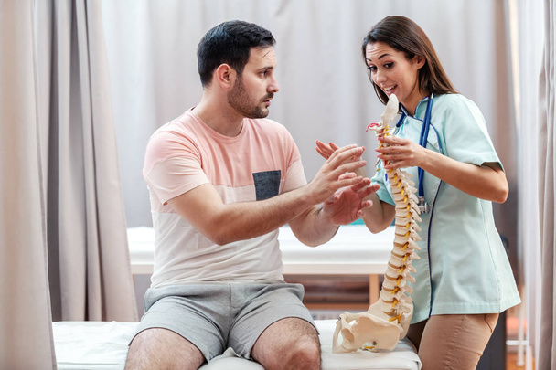 Female doctor in uniform holding spine model and talking to patient while patient showing her where he feeling pain. - Photo, Image