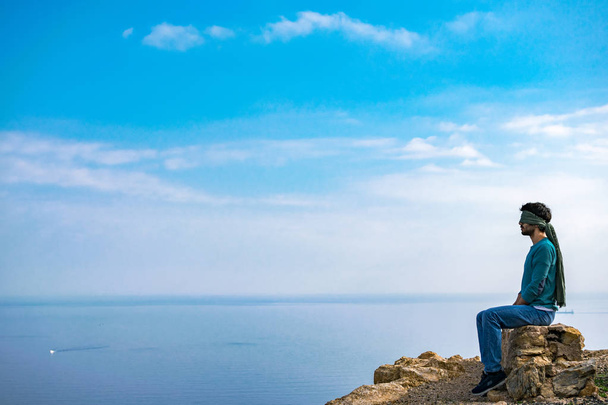 Молодой человек с повязкой на глазах на вершине утёса над морским пейзажем
 - Фото, изображение