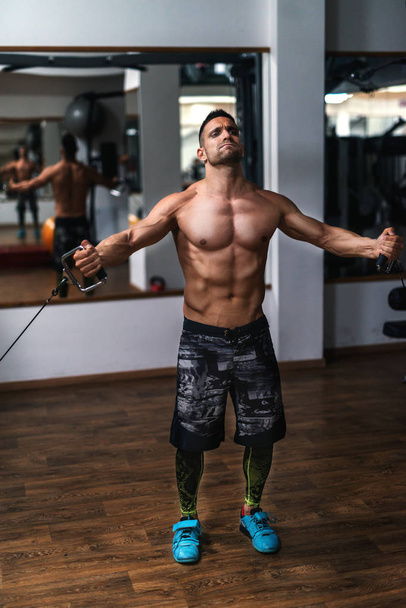 Strong shirtless bodybuilder doing exercises with exercise equipment. Gym interior. - Photo, Image