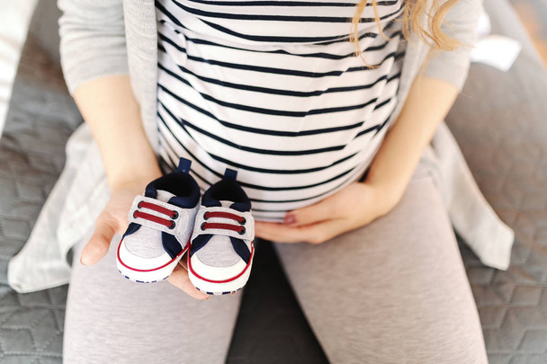 Primer plano de la mujer caucásica embarazada sosteniendo zapatos de bebé y tocando el vientre
. - Foto, imagen