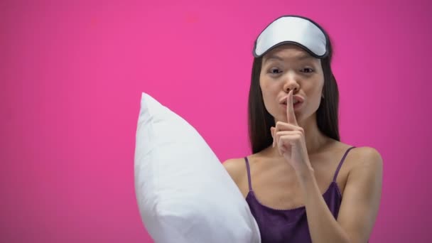 Asian lady with pillow showing silence sign and wearing eye mask, ready to sleep - Záběry, video