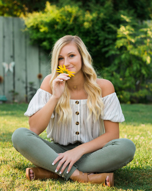 One attractive blonde hair caucasian high school senior posing for senior pictures. Female Teenager portrait outdoors at park holding yellow flower in summer. - Fotografie, Obrázek