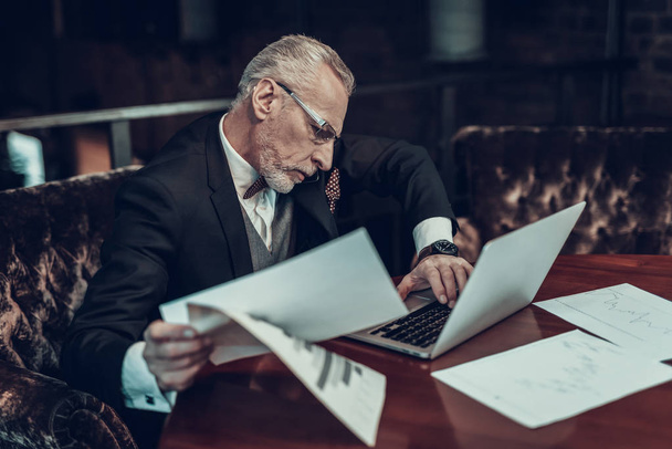 Businessman Using Laptop. Old Businessman . Man is Wearing in Black Suit. Experienced Entrepreneur. Strict Man. looking at Laptop. Working Late. holding piece of paper. talking on Phone . - Fotografie, Obrázek