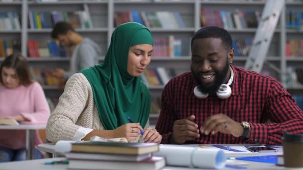 Belle femme musulmane en hijab expliquant un nouveau projet de croquis à un étudiant afro-américain positif pendant ses études à la bibliothèque avec des étudiants caucasiens flous apprenant en arrière-plan
. - Séquence, vidéo