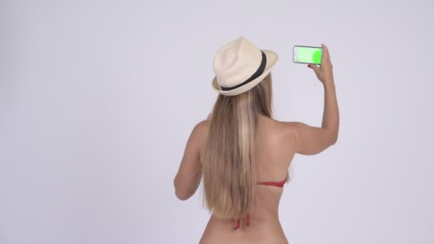 Rear view of young blonde tourist woman in bikini taking picture with phone - Πλάνα, βίντεο