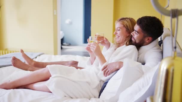 Happy mixed race young adult couple reclining on the bed in a hotel room in bathrobes holding glasses of wine - Metraje, vídeo