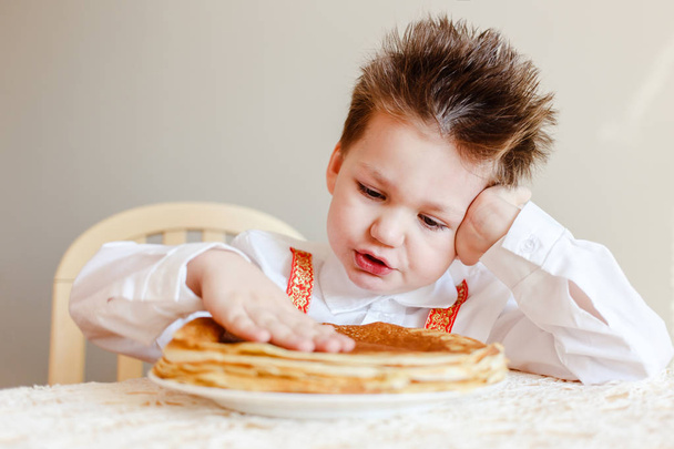 a four-year-old boy in a smart shirt touches warm pancakes with a pen. The holiday of Maslenitsa. - Zdjęcie, obraz