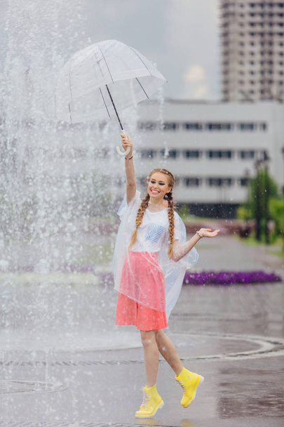 Young pretty girl with two braids in yellow boots and with transparent umbrella stands near fountain. Rainy day in city. - Photo, Image