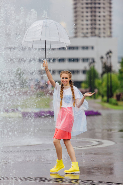Young pretty girl with two braids in yellow boots and with transparent umbrella stands near fountain. Rainy day in city. - Foto, afbeelding