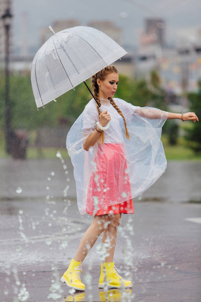 junges hübsches Mädchen mit zwei Zöpfen in gelben Stiefeln und mit transparentem Regenschirm steht in der Nähe des Brunnens. Regentag in der Stadt. - Foto, Bild