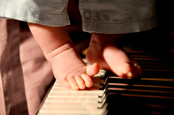 Closeup view on male babyboy bare feet with small toys playing on piano forte key board musical instrument with warm light indoor, horizontal picture - Photo, Image