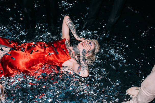 drunk woman covering face while lying on floor with confetti near people on party isolated on black - Фото, изображение