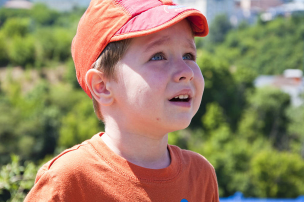 lindo niño pequeño o niño con la cara llorando en gorra roja y camisa naranja al aire libre soleado en el fondo de árboles verdes naturales en verano
 - Foto, Imagen