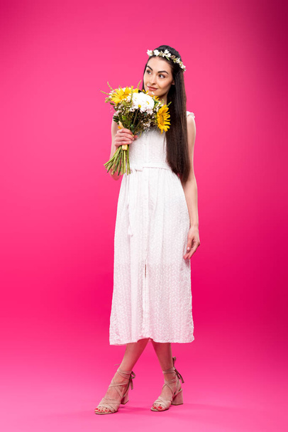 full length view of happy young woman in white dress holding flower bouquet and looking away on pink - Photo, image