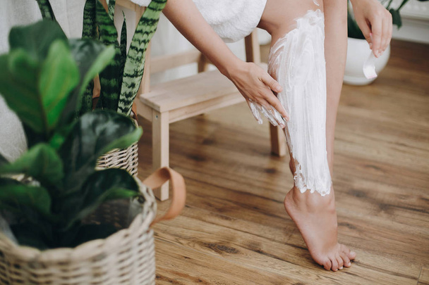 Young woman in white towel applying shaving cream on her legs in home bathroom with green plants. Skin care and wellness concept. Hand smearing moisturizer cream on skin - Fotó, kép