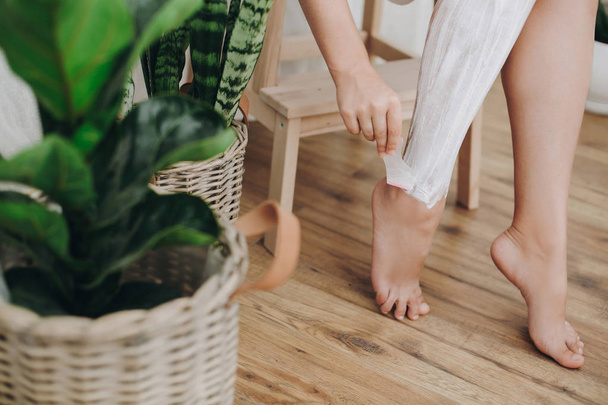 Молодая женщина в белом полотенце наносит крем для бритья на ноги в домашней ванной комнате с зелеными растениями. Уход за кожей и оздоровительная концепция. Рука держит пластиковую бритву на коже со сливками
 - Фото, изображение