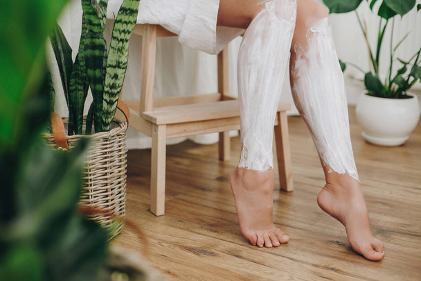 Hair Removal concept, depilation cream. Young woman in white towel applying shaving cream on her legs in home bathroom with green plants. Skin care and wellness concept. - Photo, Image
