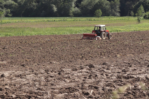 Фермер на червоному тракторі з сіячем сіє зерно в оранці землі в приватному полі в селі. Механізація весняно-польових робіт. Повсякденне життя фермера. Переробка землі. Аграрний
 - Фото, зображення