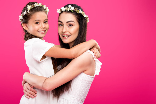 vista lateral de la hermosa madre feliz y la hija en vestidos blancos y coronas florales abrazando y sonriendo a la cámara aislada en rosa
 - Foto, imagen
