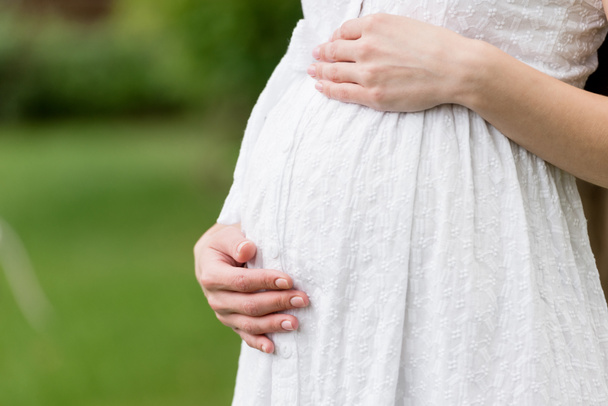 частичный вид беременной женщины в белом платье, касающейся живота, стоя в парке
  - Фото, изображение