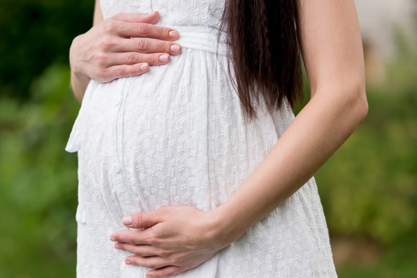 mid section of pregnant woman in white dress touching belly while standing in park  - Foto, Imagem