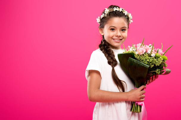 szczęśliwe dziecko w białej sukni i wieniec kwiatowy trzymając bukiet kwiatów i uśmiecha się do kamery na różowym tle   - Zdjęcie, obraz