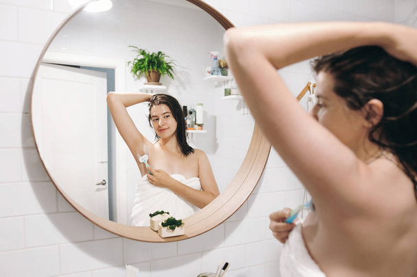 Mujer atractiva joven en toallas blancas axilas de afeitar, mirando en el espejo en el baño con estilo. Cuidado de la piel y el cuerpo. Concepto de depilación. Mujer después de afeitarse la ducha con afeitadora
 - Foto, imagen