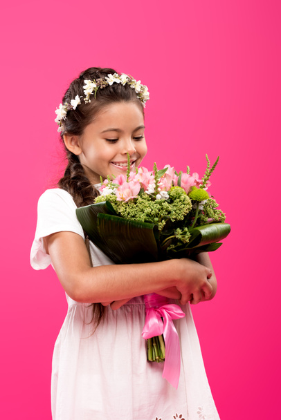adorable happy child in white dress and floral wreath holding flower bouquet isolated on pink  - Foto, Imagem