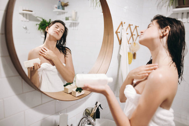 Relaxed sexy woman in white towel applying moisturizing cream on shoulder in bathroom, mirror reflection. Skin and body care. Slim young woman enjoying spa and wellness - Photo, Image