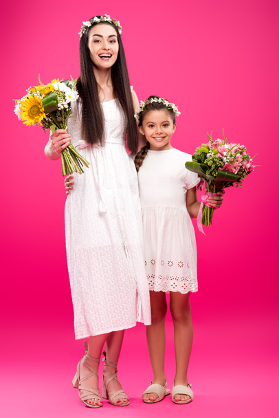 happy mother and daughter in white dresses and floral wreaths holding beautiful bouquets on pink - Foto, Imagem