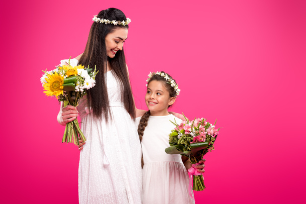 beautiful happy mother and daughter in white dresses and floral wreaths holding bouquets isolated on pink - Foto, Imagem