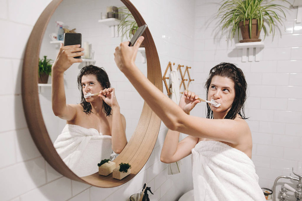 Young happy woman in white towel making selfie while brushing teeth and looking at phone in stylish bathroom at round mirror. Social media affect. Tooth care concept - Foto, Bild