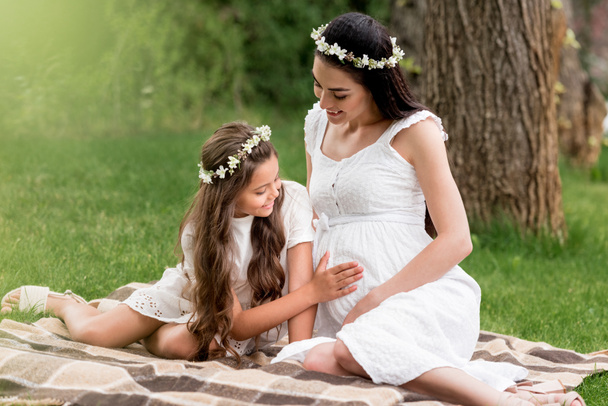 cute smiling daughter touching belly of pregnant mother while sitting together on plaid in park - Photo, Image