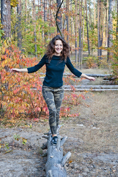 young sexy woman or girl with long hair on pretty smiling face in black shirt and camouflage jeans walks on tree trunk in autumn seasonal forest or wood with colorful leaves on natural background - Photo, Image