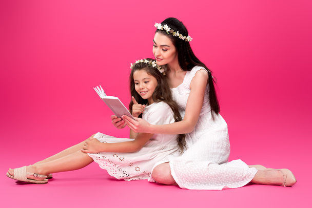 beautiful happy mother and daughter in white dresses sitting and reading book together on pink - Foto, Bild