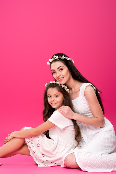 beautiful happy mother and daughter in white dresses sitting together and smiling at camera on pink - Photo, Image