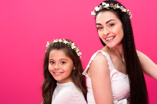 hermosa madre feliz e hija en vestidos blancos y guirnaldas florales sonriendo a la cámara aislada en rosa
 - Foto, imagen