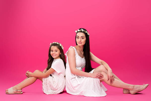 beautiful mother and daughter in white dresses sitting back to back and smiling at camera on pink - Foto, Imagem