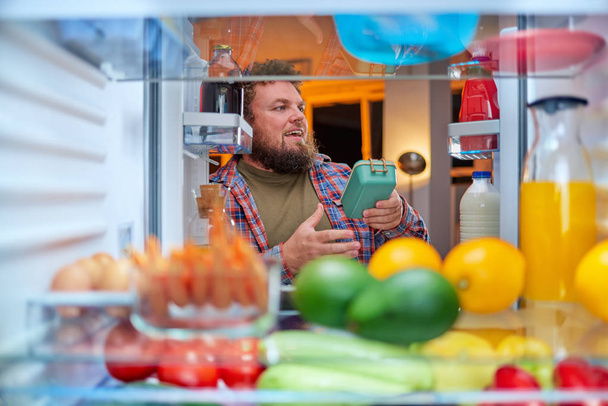 Mies etsii jotain syötävää yöllä seistessään jääkaapin edessä. Epäterveellistä syömistä. Kuva otettu jääkaapin sisältä
. - Valokuva, kuva