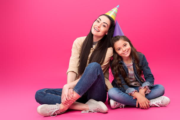 full length view of happy mother and daughter in party hats sitting together and smiling at camera on pink - Foto, imagen