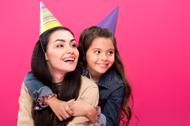 happy mother and daughter in party hats hugging and looking away isolated on pink - Photo, Image