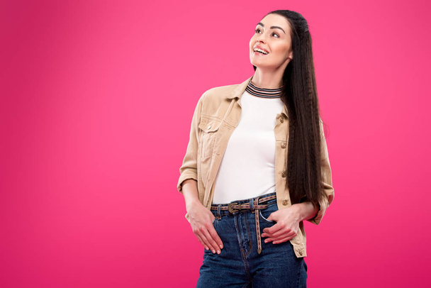smiling young woman standing with hands in pockets and looking away isolated on pink - Foto, afbeelding