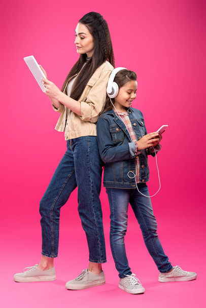 vista lateral de la madre y la hija sonrientes en auriculares de pie espalda con espalda y el uso de dispositivos digitales en rosa
  - Foto, imagen