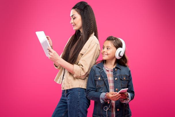 smiling daughter in headphones holding smartphone and looking at mother using digital tablet isolated on pink - 写真・画像